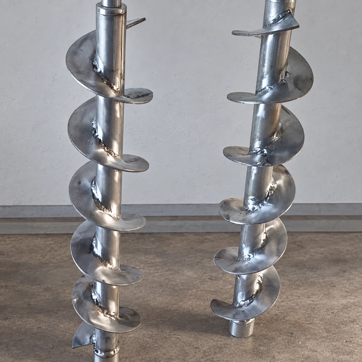 Two metal shafts with helical screw flights standing on a workshop bench.